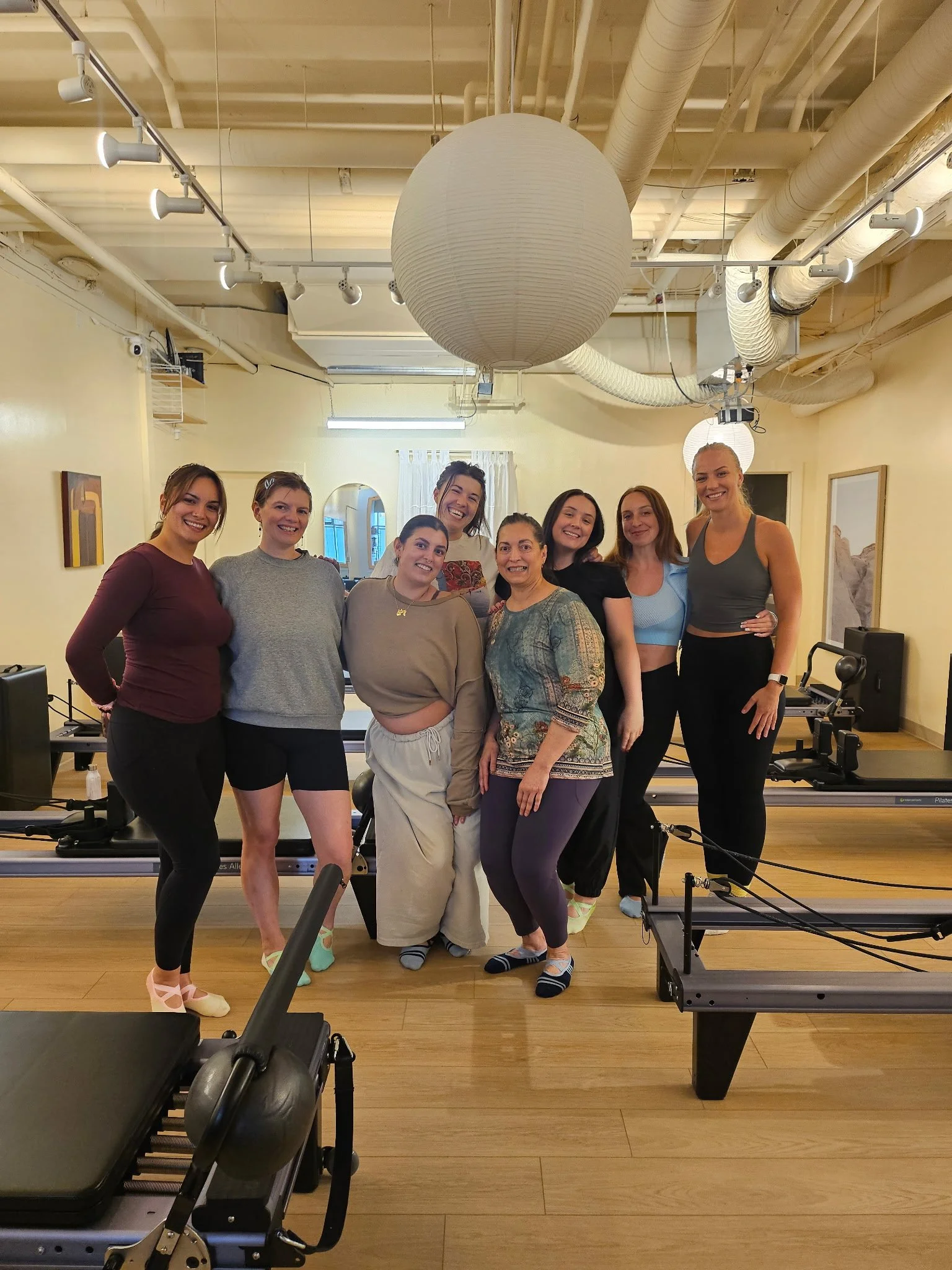 A group of nine women standing in a Pilates studio, smiling at the camera. The studio has light-colored walls, wooden floors, and Pilates reformer machines visible in the background. Large paper lanterns hang from the ceiling, and the women are dressed in athletic wear.