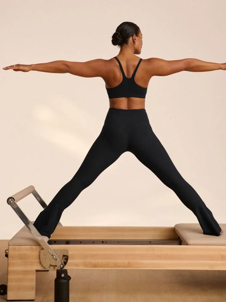 A woman in black sportswear practicing Pilates on a Reformer machine with arms extended outward.