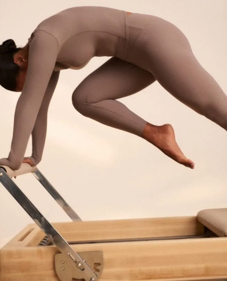 Person in beige athletic wear performing a yoga pose on a reformer machine, with hands on the platform and body supported by a ladder and the machine.