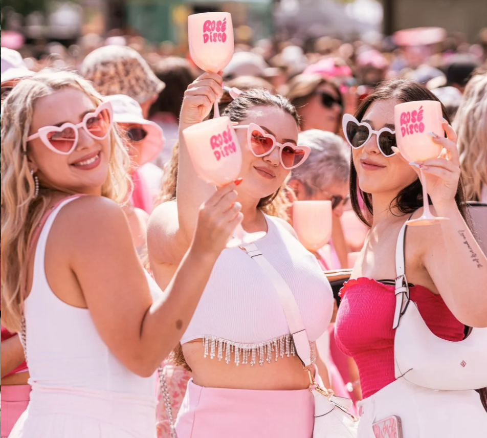 Three women at a crowded outdoor party, smiling and holding pink glasses labeled 'Rosé Disco,' wearing stylish sunglasses and summer clothes.