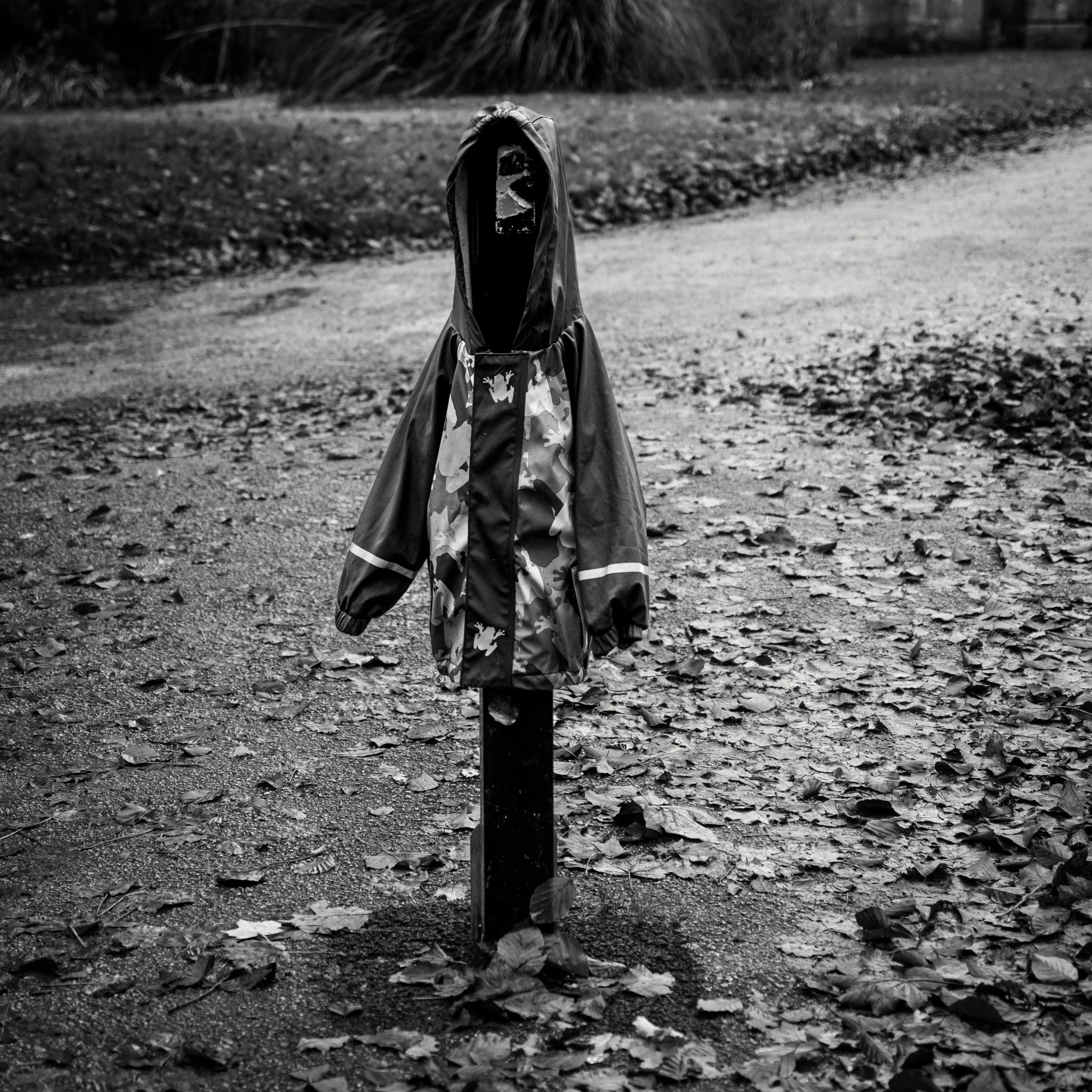 Black and white image of a child's rain jacket left on a bollard among autumn leaves.
