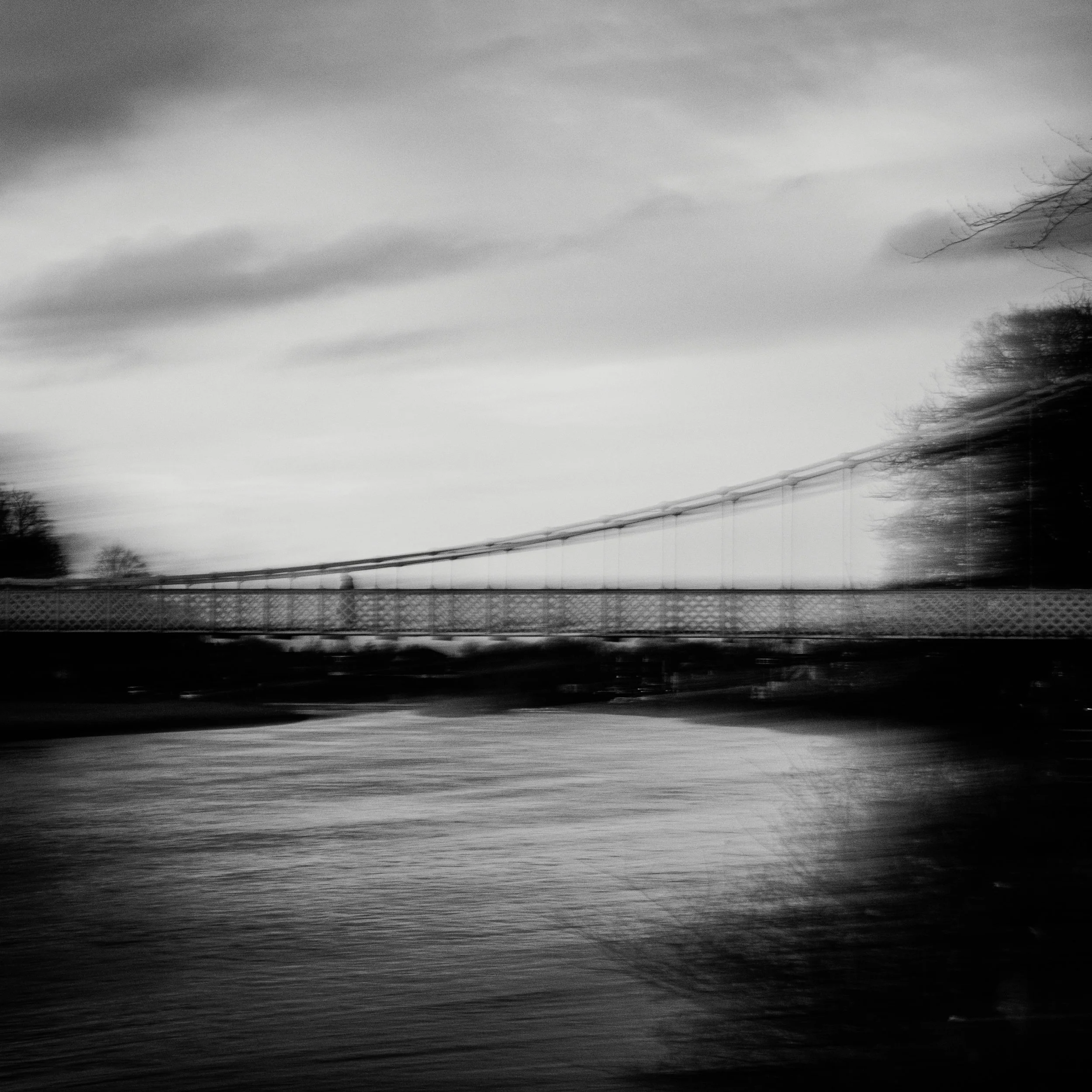 Fine art photography of a suspension bridge and river captured in moody black and white motion blur.