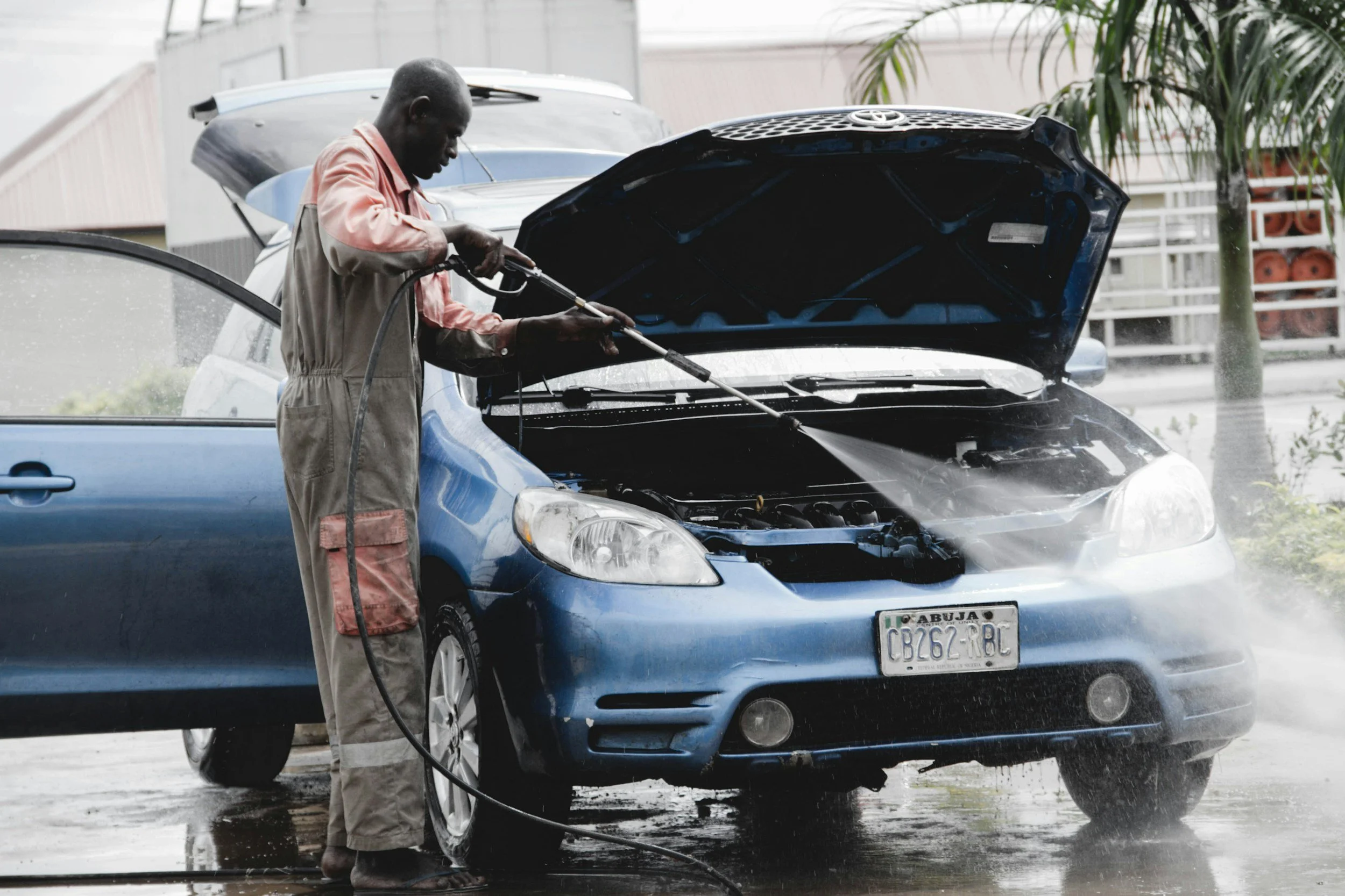 Un homme lave une voiture bleue sous la pluie, le capot ouvert.