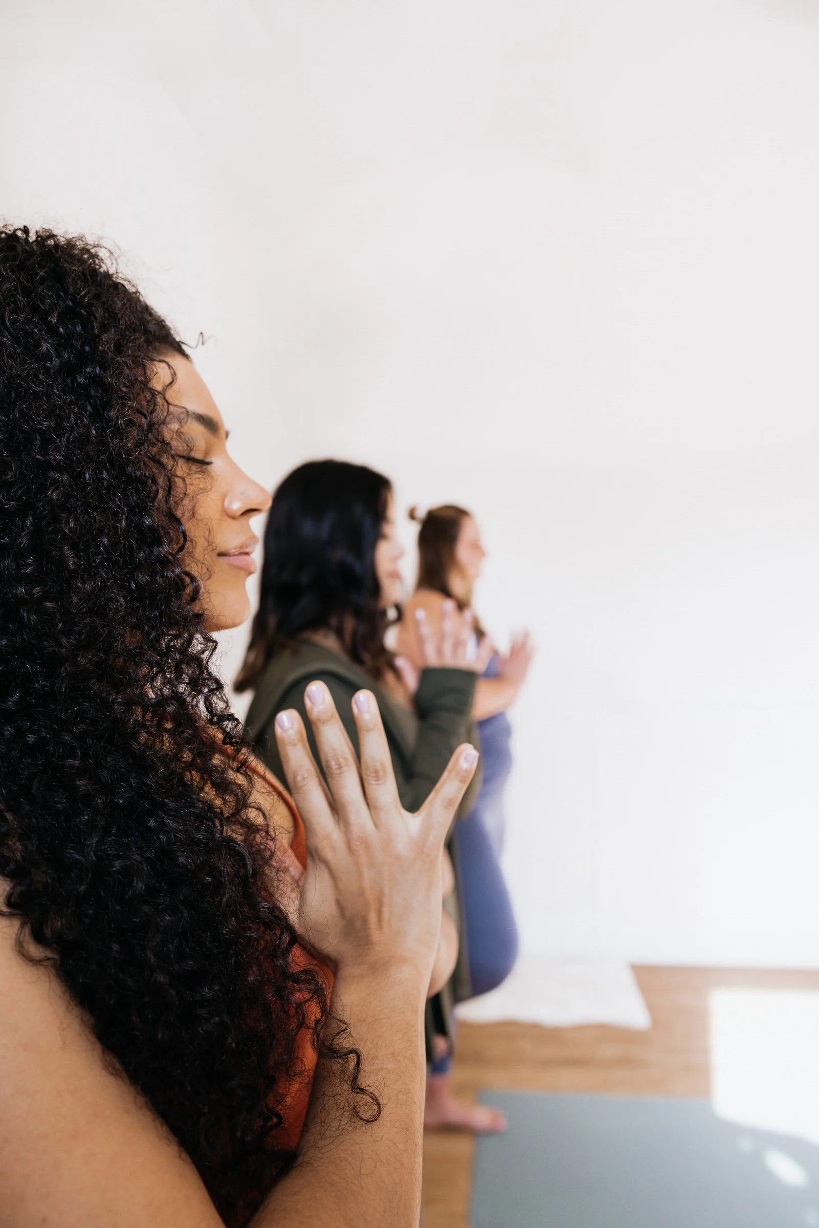 Group of women practicing yoga with eyes closed, standing in a row, in a studio with a plain white wall.