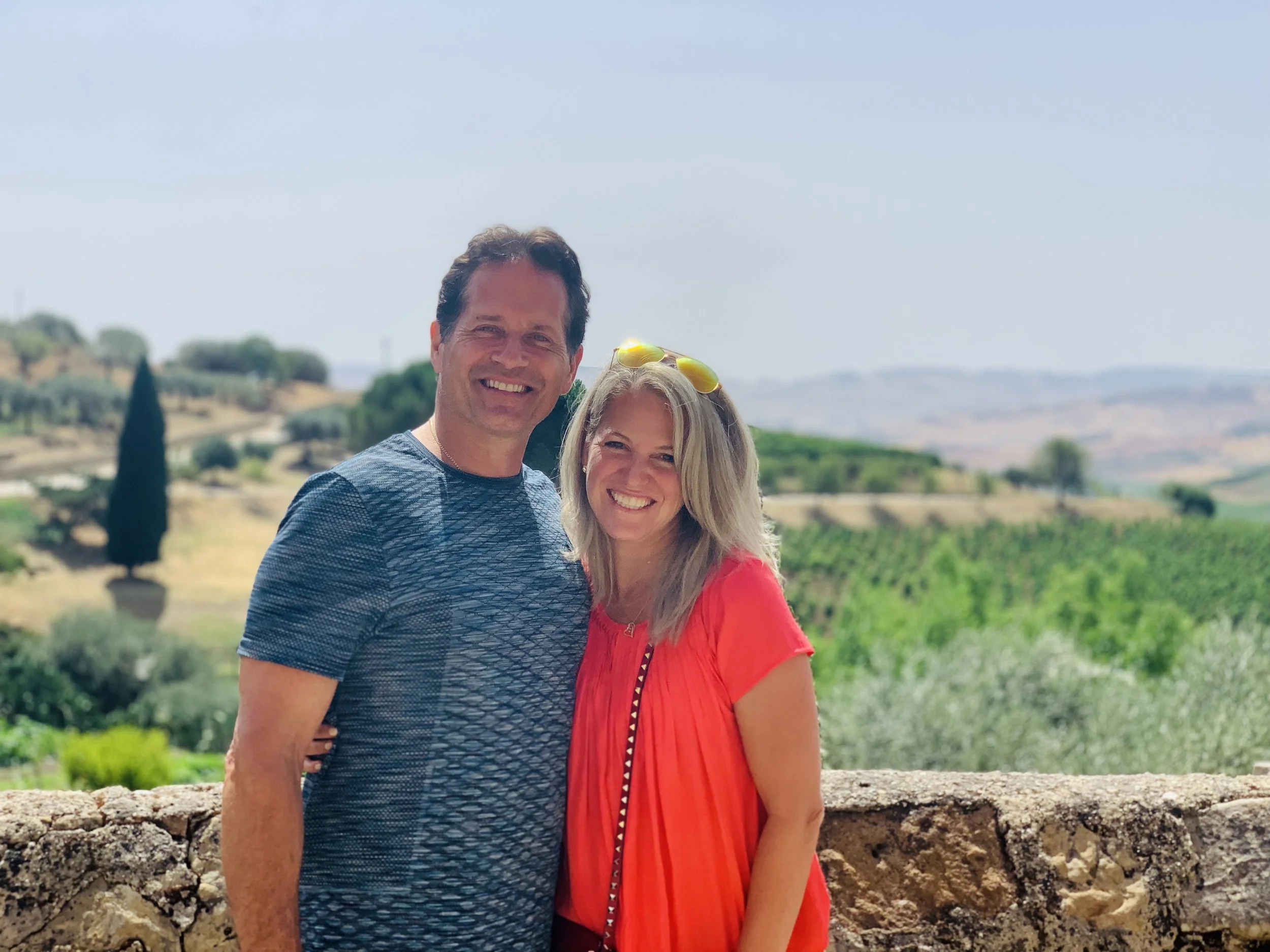 A man and woman smiling, standing outdoors in front of rolling vineyards and green landscape under a blue sky.