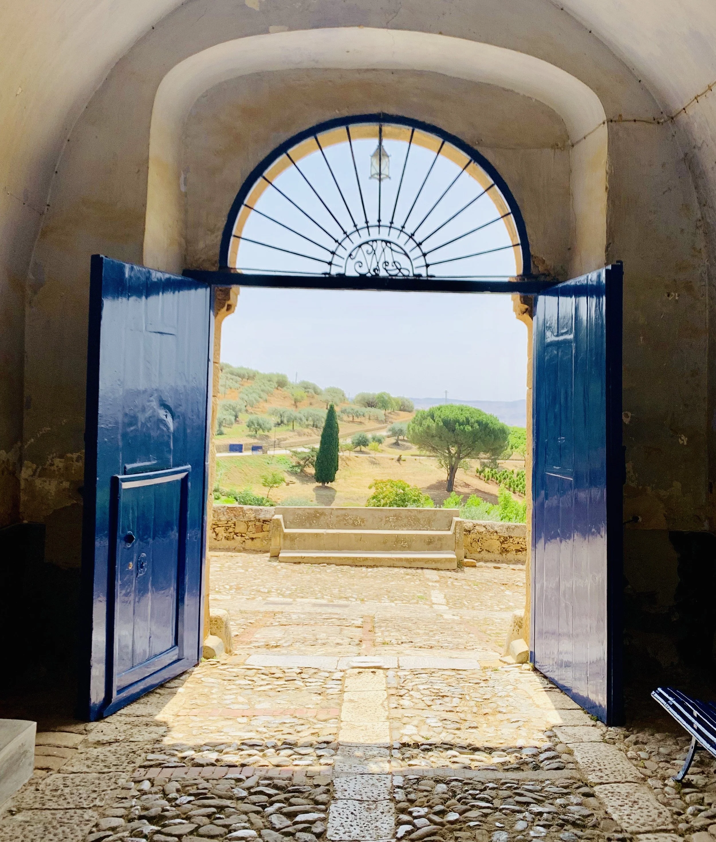 Open blue doors leading to a courtyard with a cobblestone path, view of hills and trees in the distance under a blue sky.