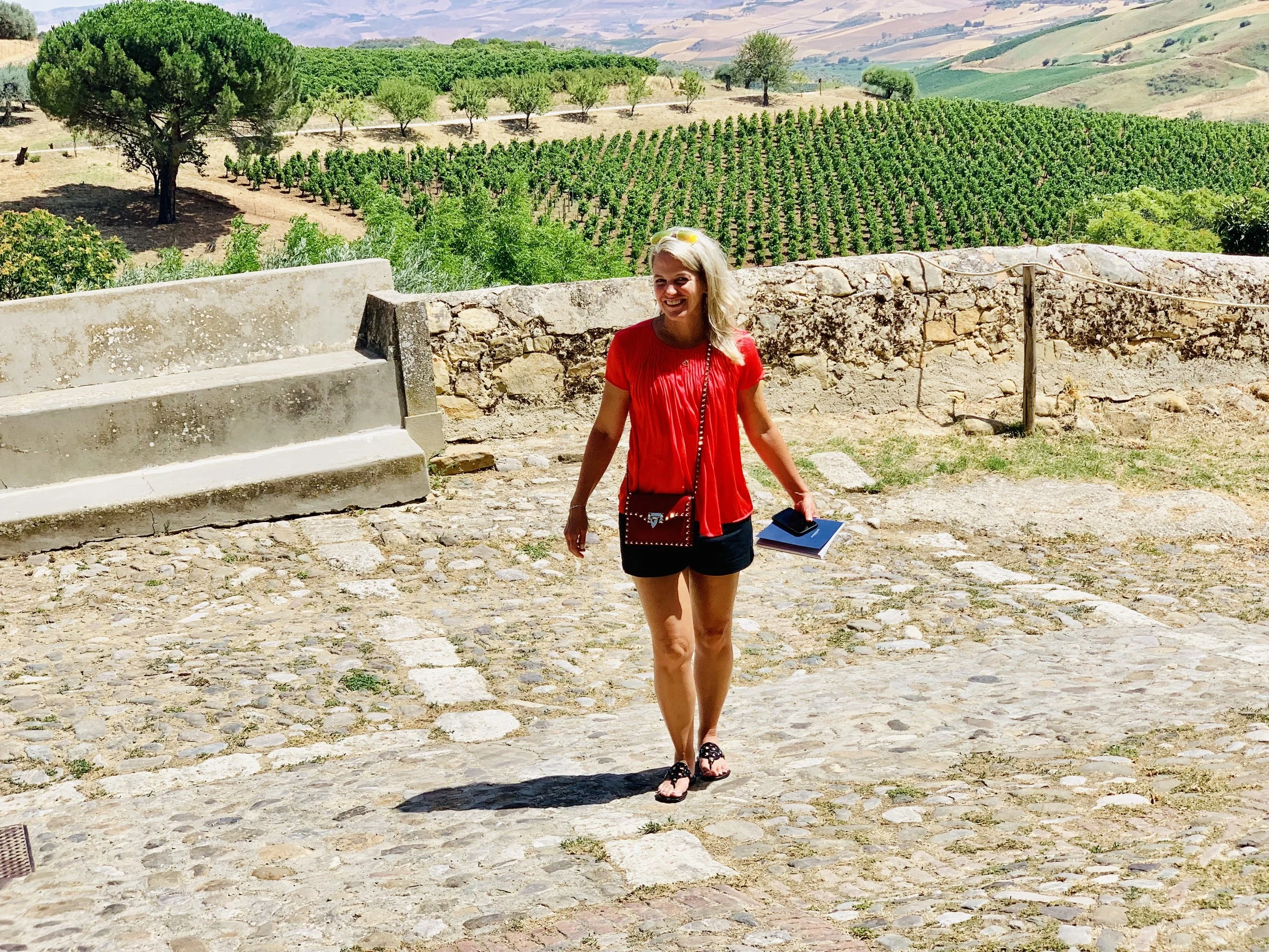 A woman in a red top and black shorts walking on a cobblestone path outdoors with vineyards and rolling hills in the background.