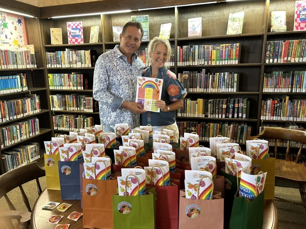 Two people smiling and holding a colorful sheet of paper in a library with bookshelves in the background. There is a table in front with multiple gift bags decorated with rainbows and animals, and some small round stickers or badges lying on the table.