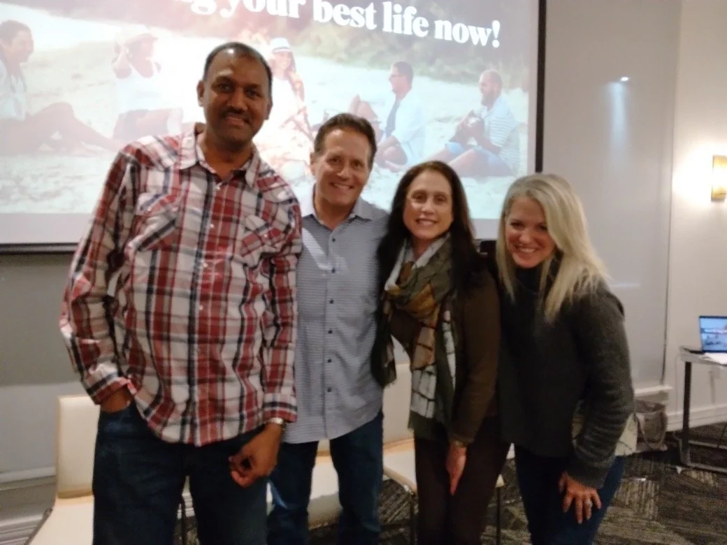 Four people standing together in a room with a presentation screen behind them showing a beach scene and the text 'your best life now!'
