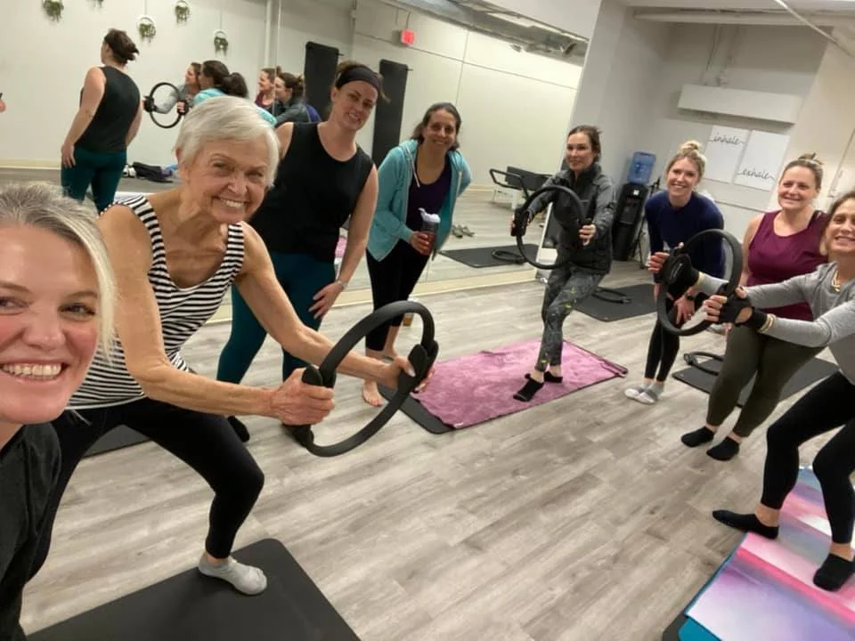 A group of women in workout attire in a fitness class, some holding exercise ring props and others holding weights, in a gym room.