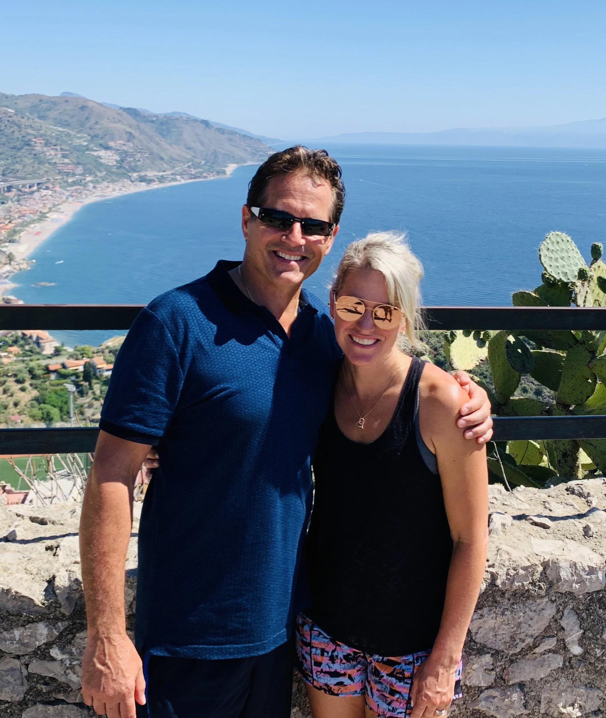 Smiling man wearing sunglasses and blue shirt, standing next to a woman in black top and patterned shorts, overlooking a coastal landscape with mountains, water, and buildings in the distance.