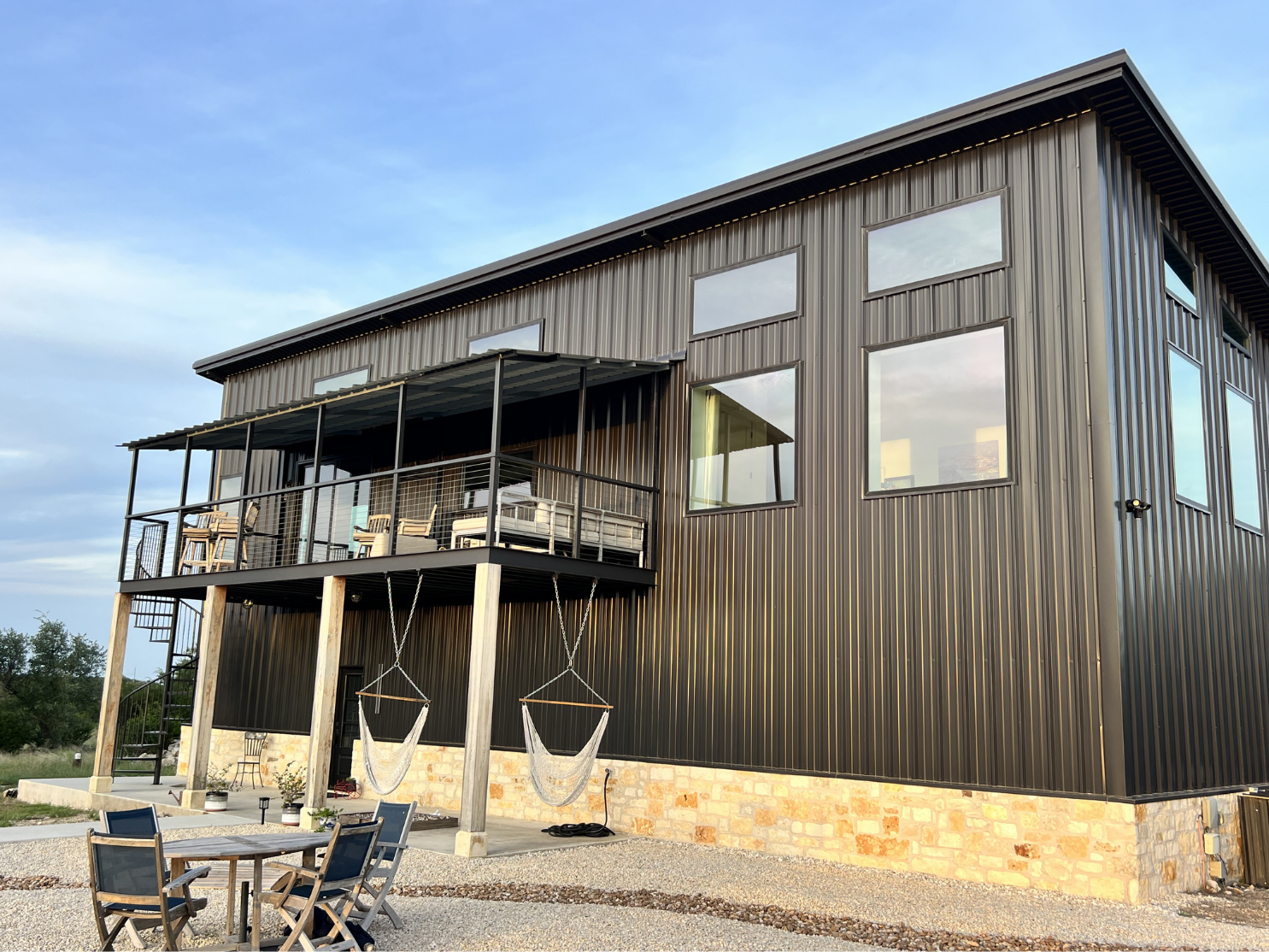 A modern black metal house with large windows, a balcony with outdoor furniture, hanging hammock chairs, and a gravel patio with a wooden table and chairs.