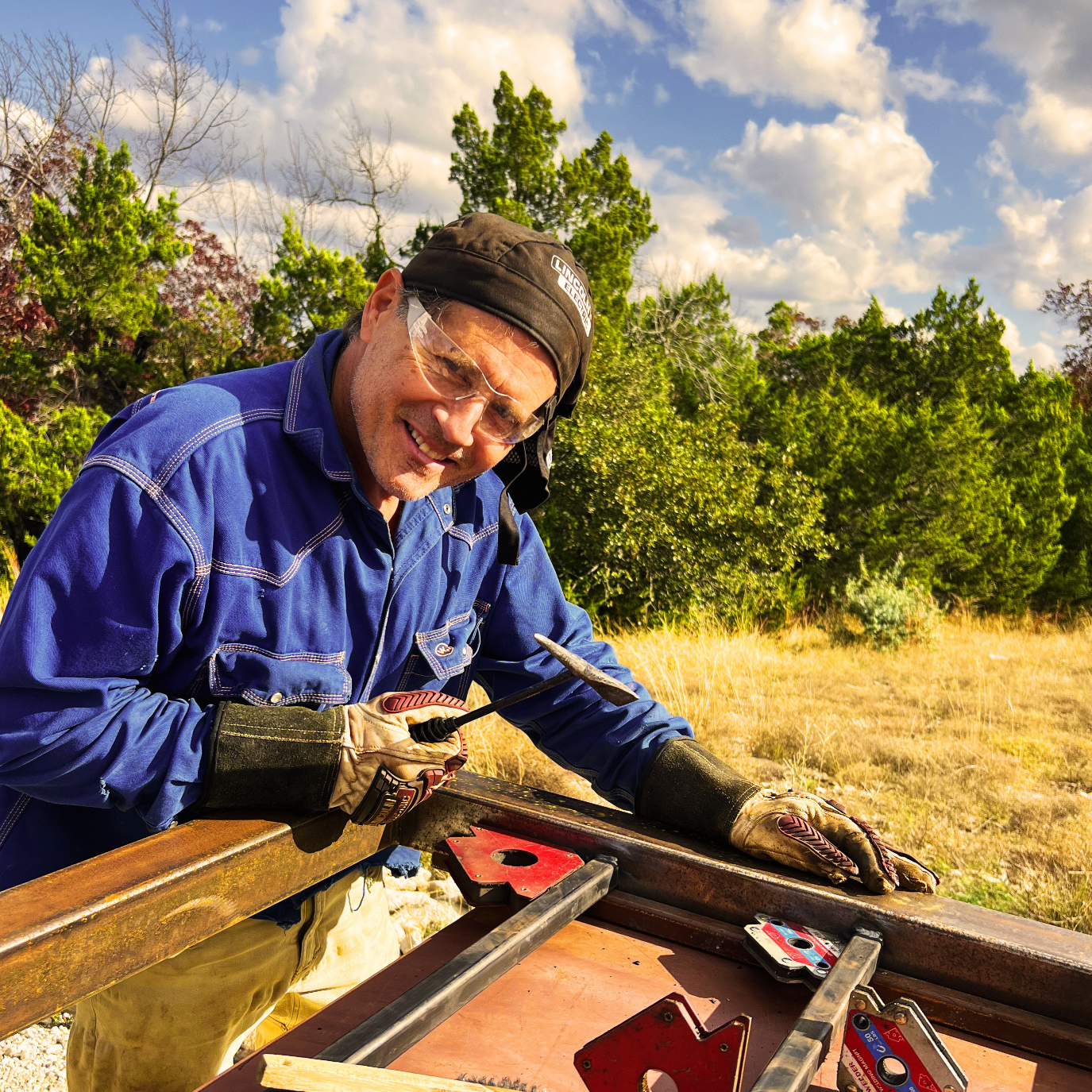 A man in safety glasses, a black cap, a blue work shirt, and gloves is working with metal outdoors during daytime. There are trees and clouds in the background.