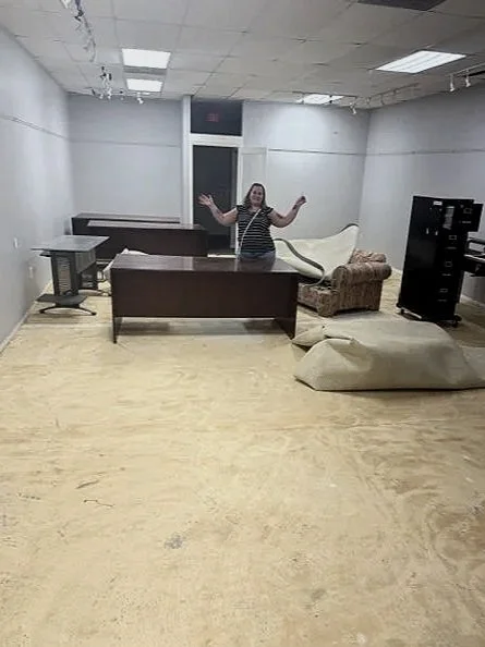 A woman standing in a mostly empty room with some furniture, including desks, a sofa, a beanbag, and a filing cabinet, with her arms raised. celebrating my new space for thumbprint art cafe