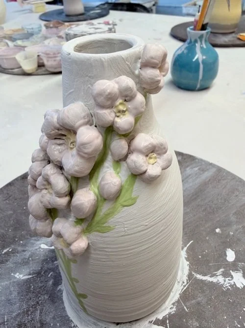 A ceramic vase with a textured surface, decorated with 3D pastel pink and purple flower blossoms and green stems, placed on a worktable surrounded by art supplies. made it all by myself, made with pottery wheel and sculpting flowers