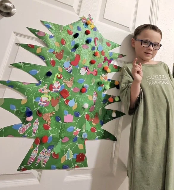 Child standing next to a large, decorated paper Christmas tree on a door. Little Hands Activity