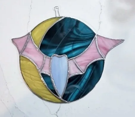 Try stained glass for the first time or use our tools to create stained glass as a seasoned artist. A stained-glass art piece with abstract shapes in yellow, pink, blue, and teal colors. It is mounted on a white wall.