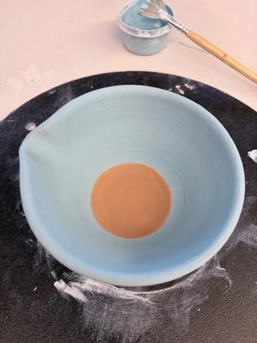 Handmade Painted ceramic bowl with a light blue exterior and a brown circular center, placed on a black table with paint splatters. A small container of blue paint and a wooden-handled paintbrush are in the background.