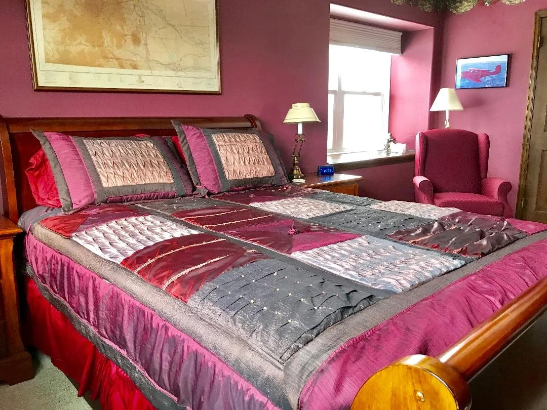 A bedroom with a large wooden bed covered with a multicolored quilt and matching pillows, pink walls, a window with a white shade, a wooden nightstand with a lamp and clock, and a pink armchair next to a wall-mounted TV.