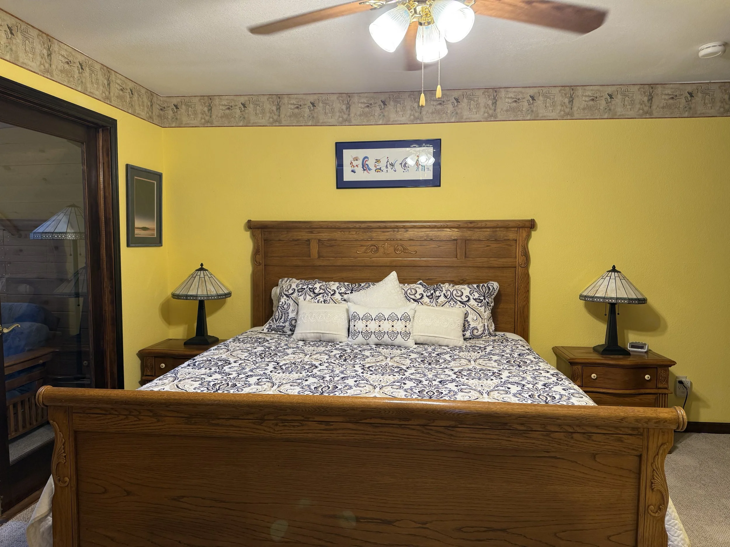 A bedroom with yellow walls, a wooden bed frame with a patterned bedspread, matching pillows, two bedside tables with lamps, a ceiling fan, and a small framed picture above the bed.
