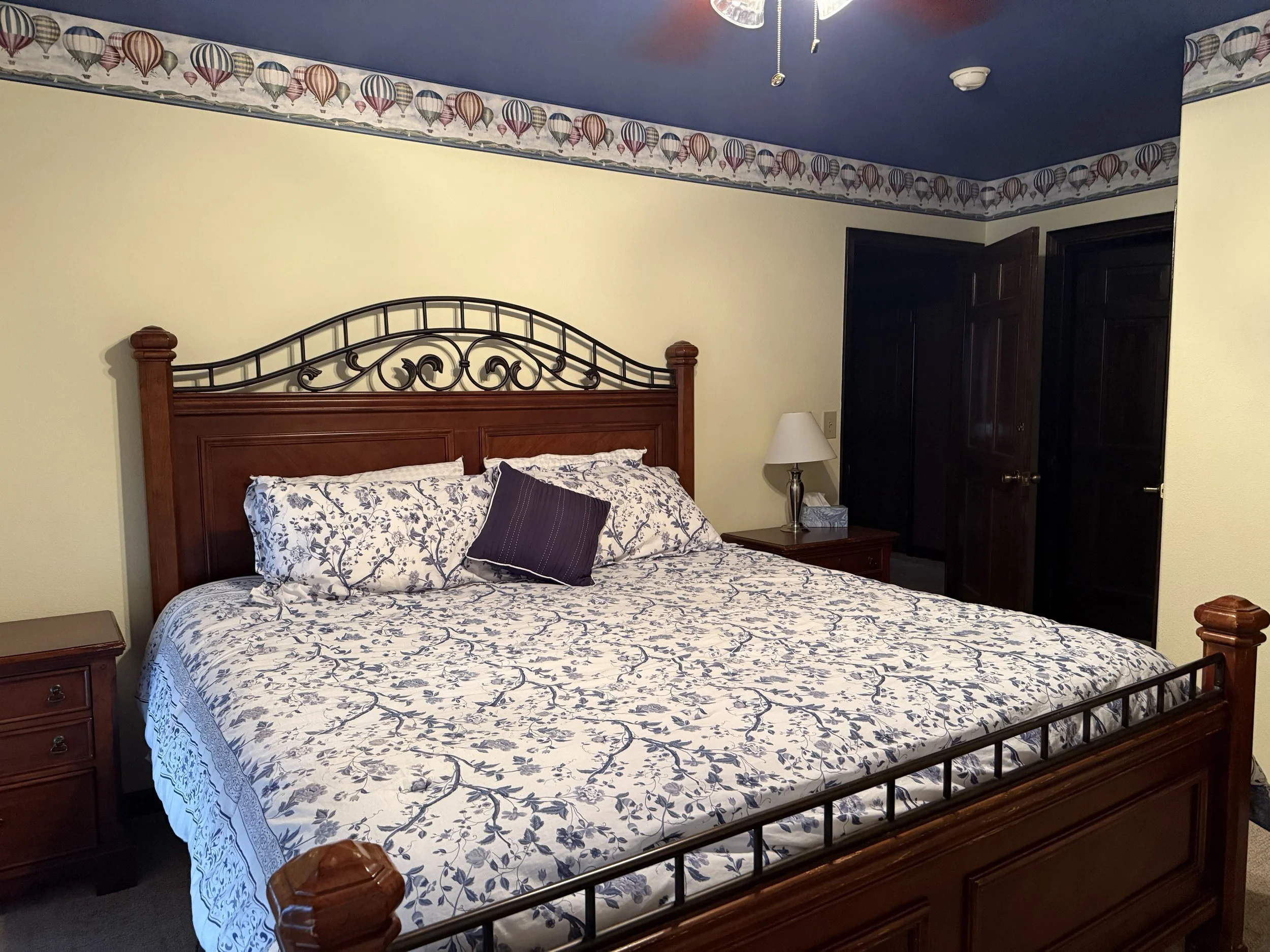 A bedroom with a wooden bed frame, white and blue floral bedding, and a purple accent pillow. There are two nightstands with lamps, dark closet doors, light yellow walls, and a border with hot air balloon patterns near the ceiling. The ceiling is painted blue.