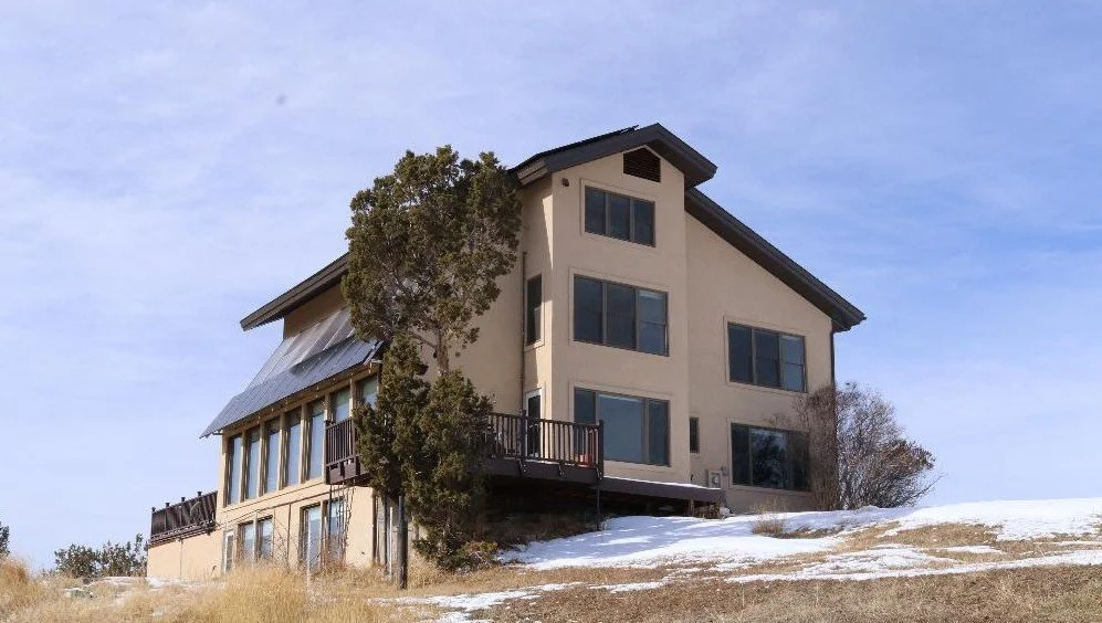 Large modern house on a hillside with snow patches, a tall tree in front, and a clear blue sky.