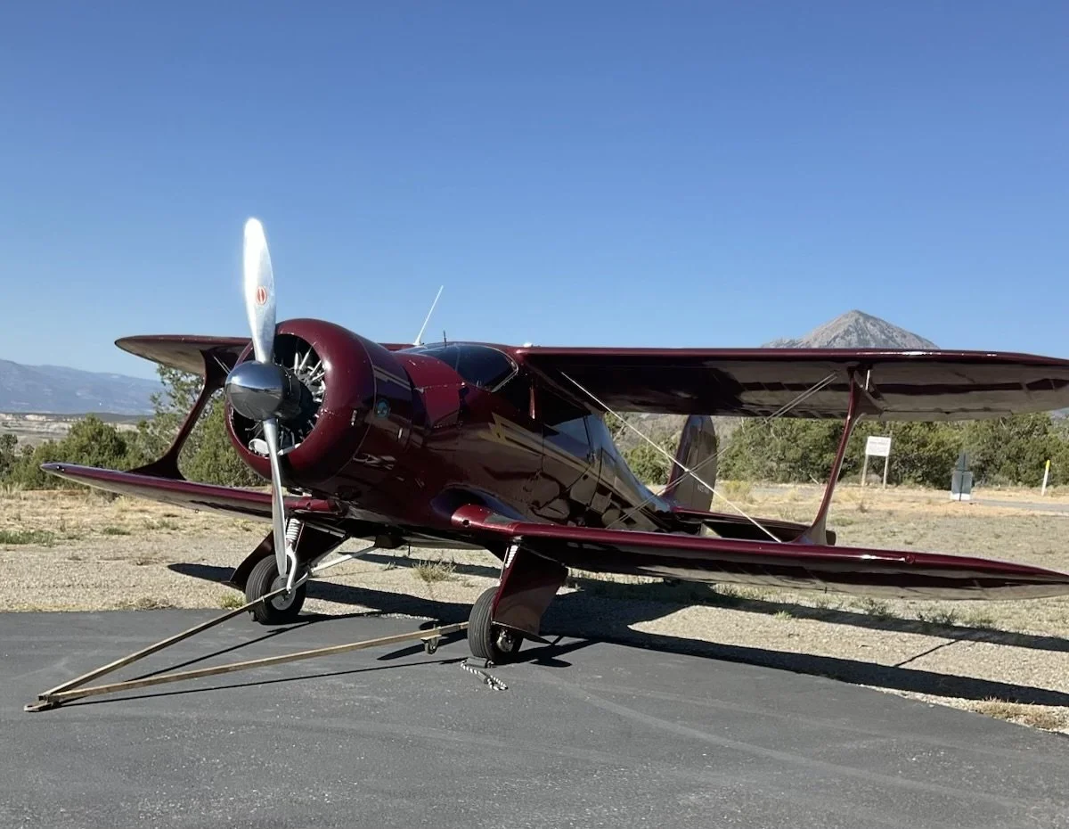 Maroon vintage airplane parked on a tarmac with mountains and blue sky in the background.