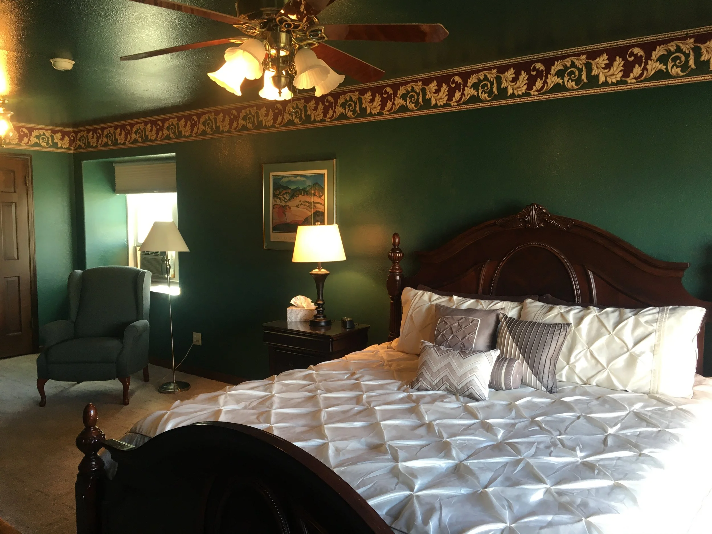 A bedroom with green walls, a wooden bed with pillows and a textured white comforter, a nightstand with a lamp, a blue armchair, a window with a shade, and decorative border on the ceiling.