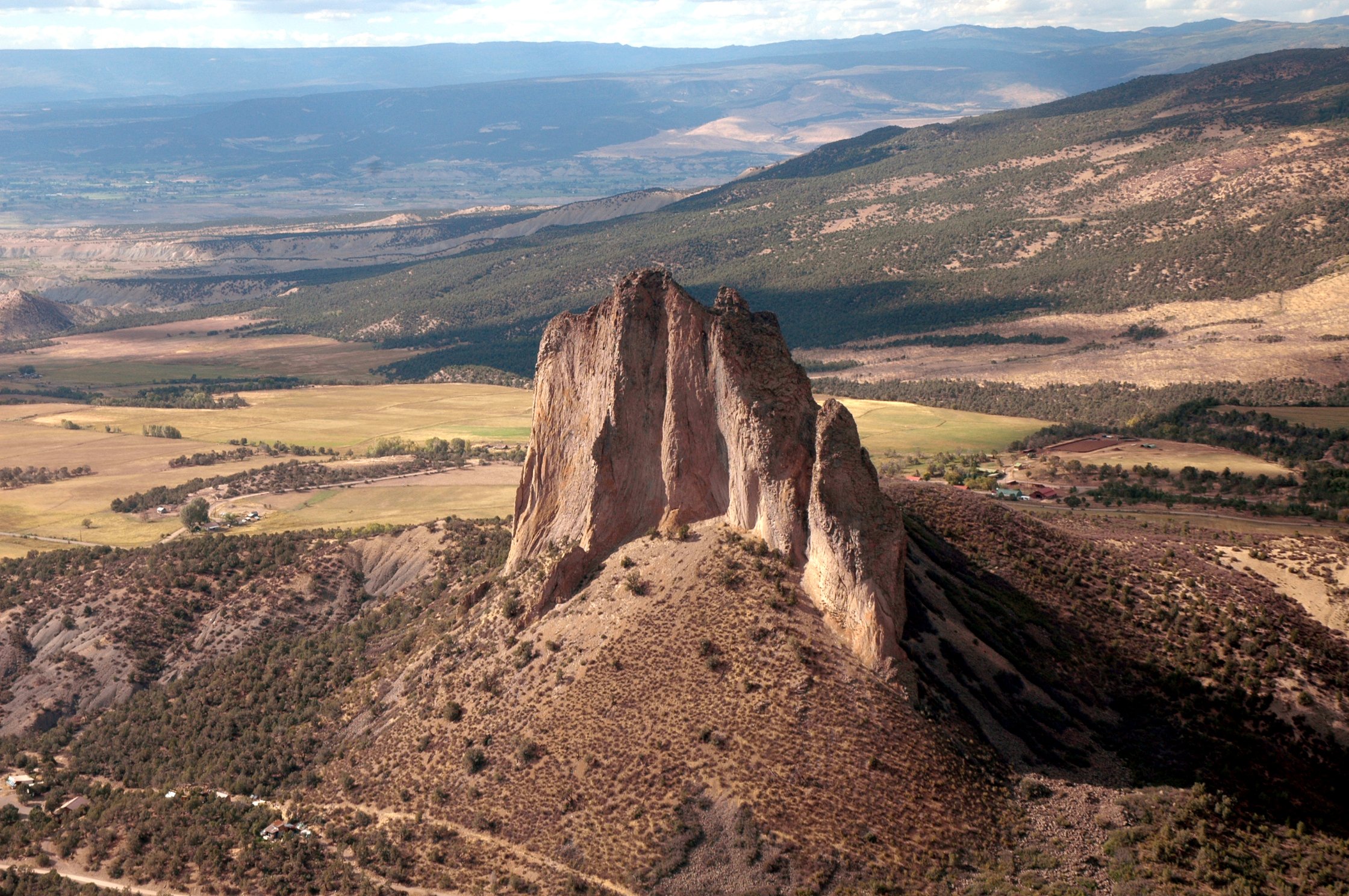 A tall, steep rock formation in a desert landscape with mountains in the background and agricultural fields at the base.
