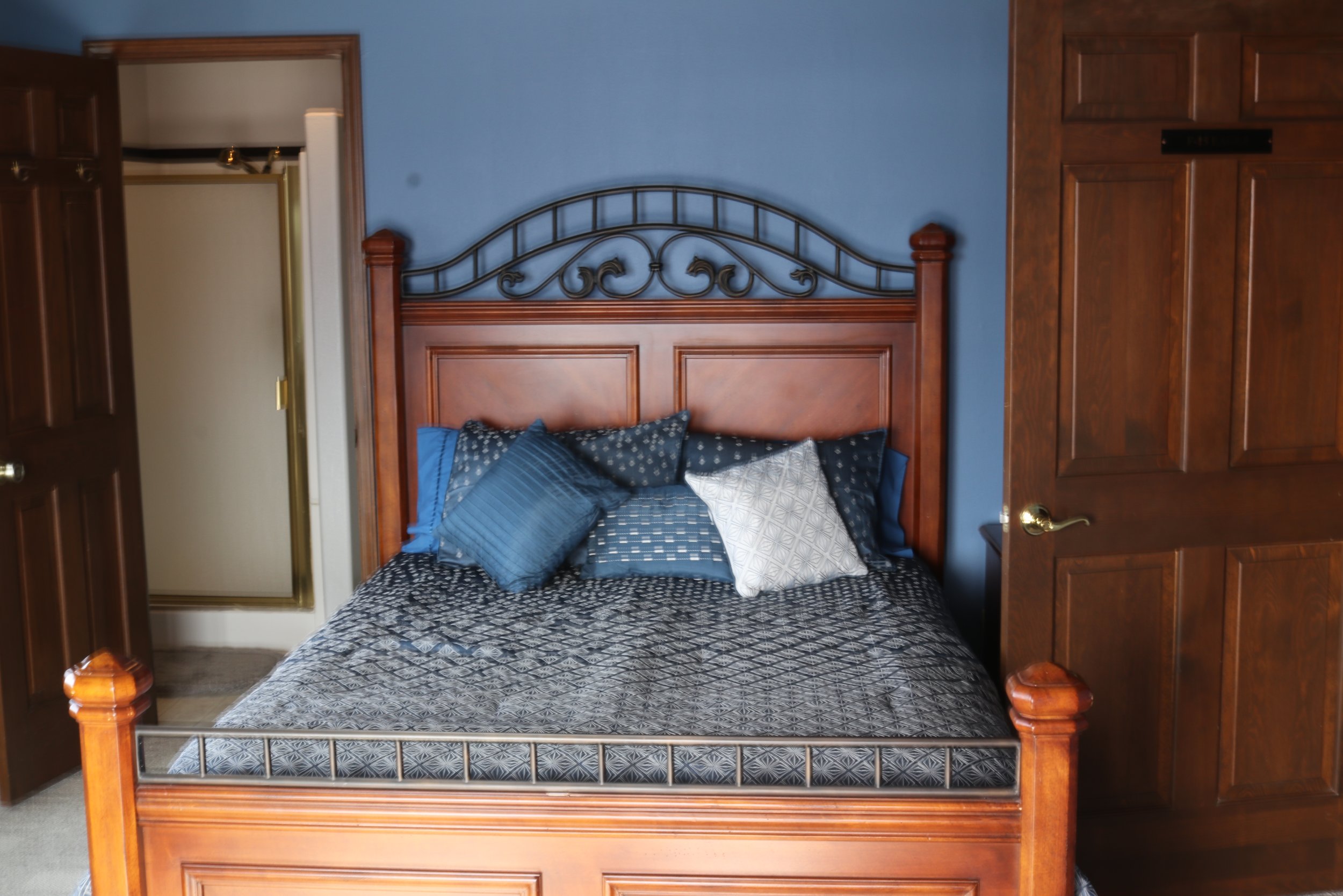A bedroom with a wooden bed frame, blue wall, and brown wooden closet. The bed has a black, gray, and white patterned bedspread with multiple blue and white pillows.