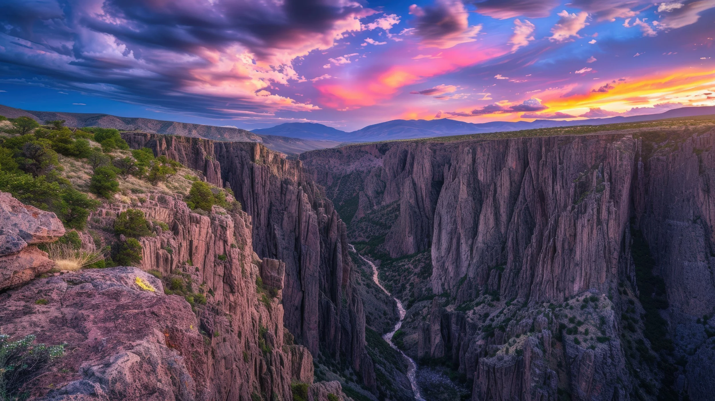 A scenic view of a deep canyon with tall, rocky cliffs and a winding river at the bottom. The sky is colorful with shades of pink, purple, and orange during sunset, and distant mountains are visible on the horizon.