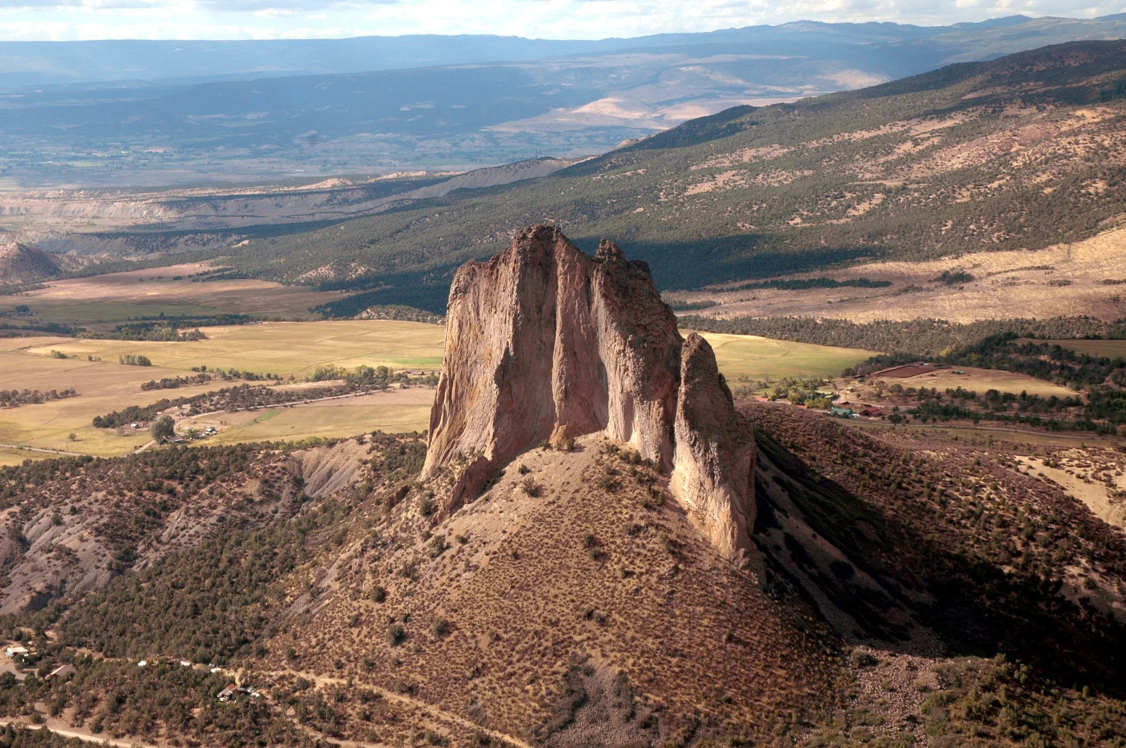 A large, jagged rock formation rising from a desert hillside with agricultural fields and mountains in the background.