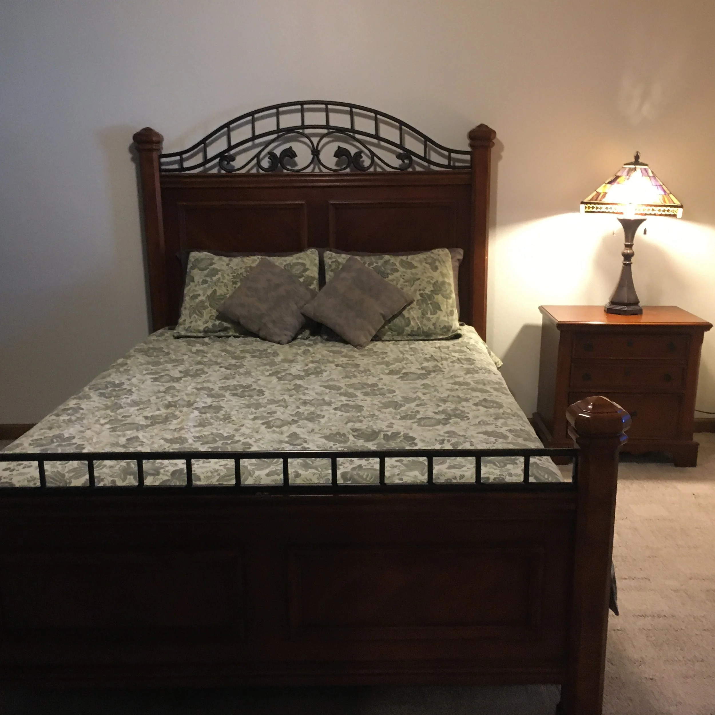 A bedroom with a wooden bed frame, patterned bedding, and a nightstand with a lamp.