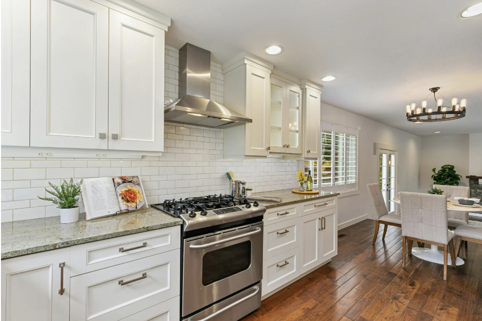 Modern kitchen with white cabinets, granite countertops, white subway tile backsplash, stainless steel stove and range hood, and hardwood floors. There are small plants and open cookbooks on the counter, with a dining area visible in the background.