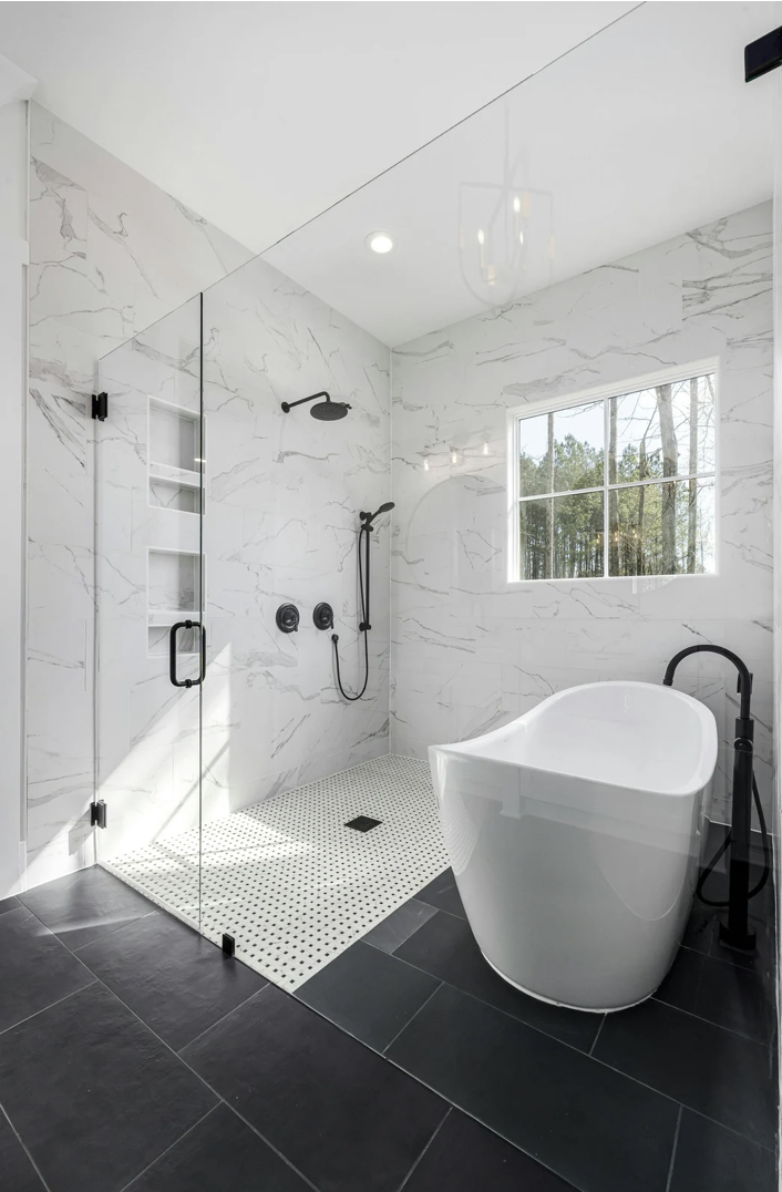 Modern white bathroom with a freestanding bathtub, a walk-in shower with black fixtures, black tiling, and a large window showing trees outside.