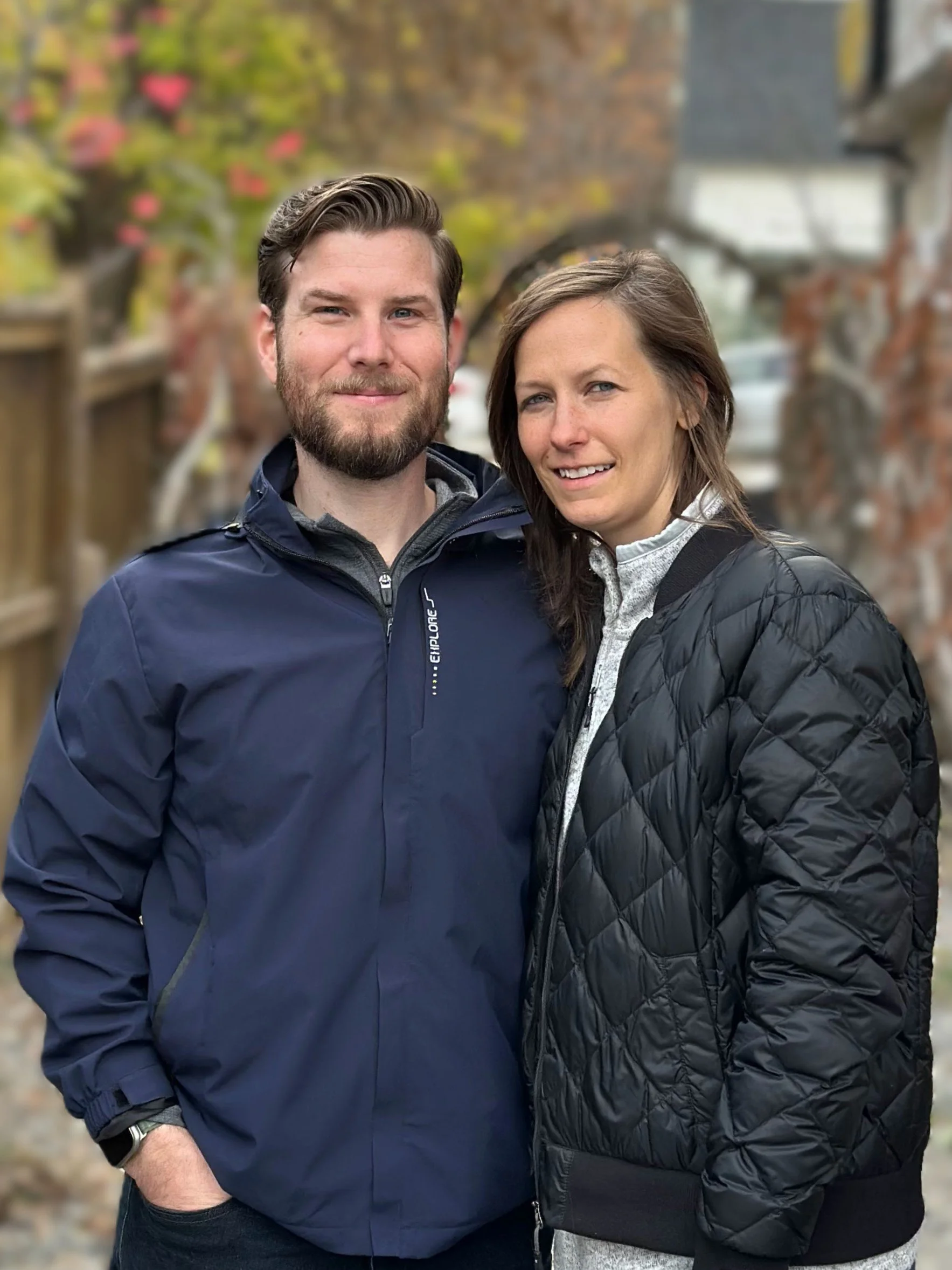 A man and woman standing outdoors on a fall day, smiling and posing for a photo. The man has short brown hair and a beard, wearing a navy blue jacket. The woman has long brown hair, wearing a black quilted jacket and a light-colored shirt, with colorful trees in the blurred background.