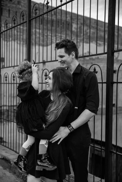 A smiling family of three, including a woman, a man, and a young girl, standing outdoors in front of a black metal fence. The woman is holding the girl, and the man is touching the woman's waist, all enjoying a happy moment together.