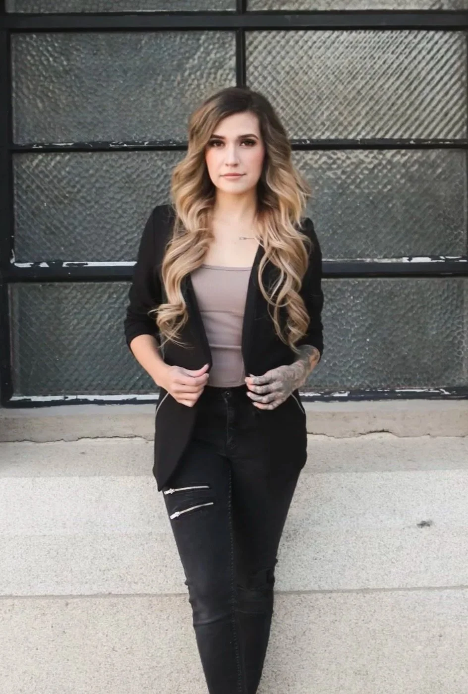 A young woman with long, wavy blonde hair standing in front of a textured glass garage door. She is wearing a black jacket, a beige top, and black jeans with zippers. She has tattoos visible on her left arm and a neutral facial expression.