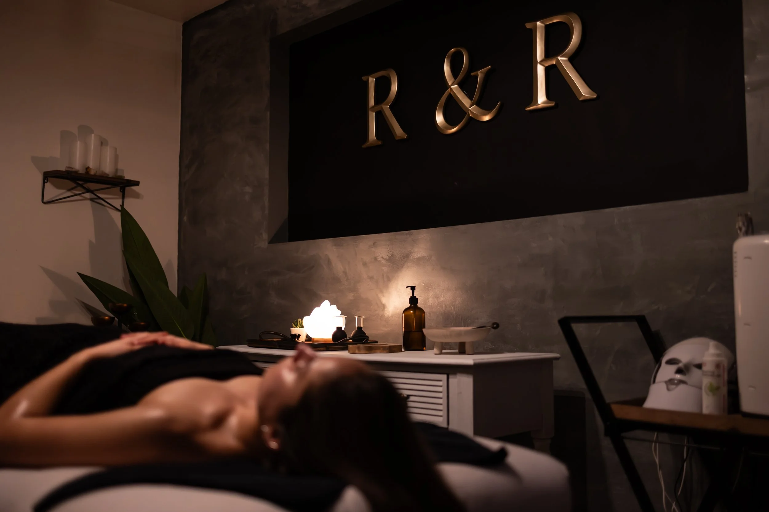A woman lying on a massage table in a dimly lit spa or massage room. The room has a black wall with large golden letters 'R & R'. There are soft candle lights, a diffuser, and decorative items on a white side table. A botanical plant is in the corner, and there is a chair with a white mask and skincare products.