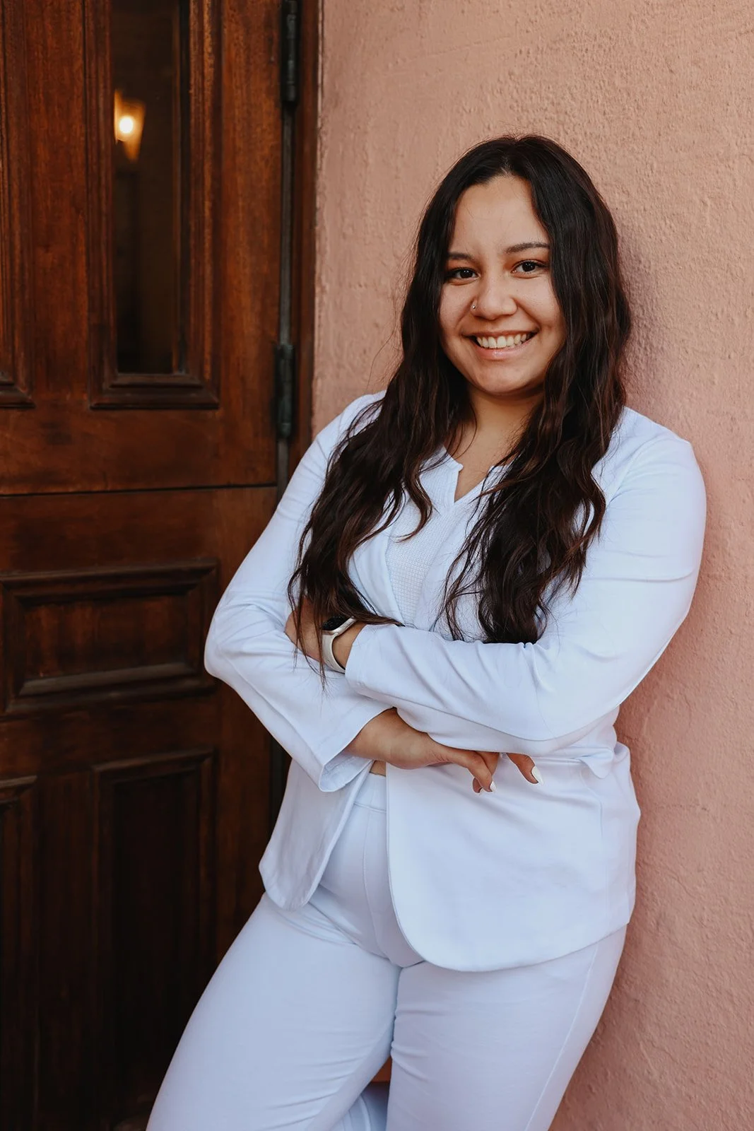 A woman with long dark hair, smiling, wearing a white blazer and pants, standing against a pink wall with a wooden door nearby.