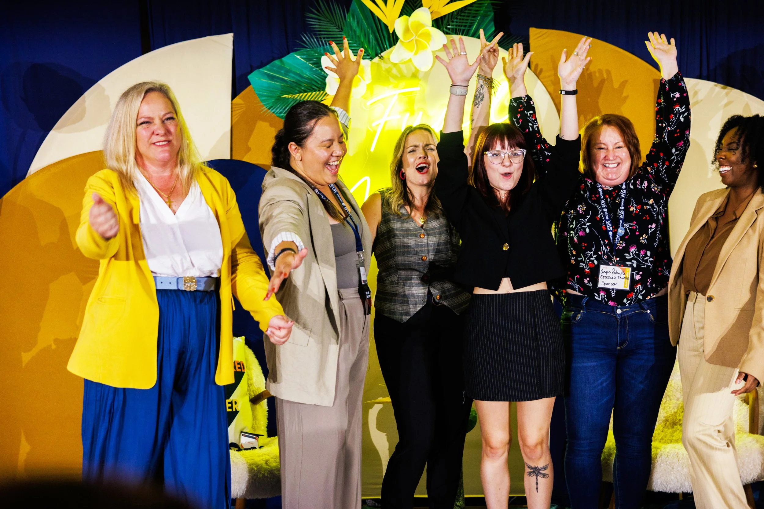 Six women on stage celebrating, some with raised hands, with colorful tropical decorations and a yellow sign in the background.