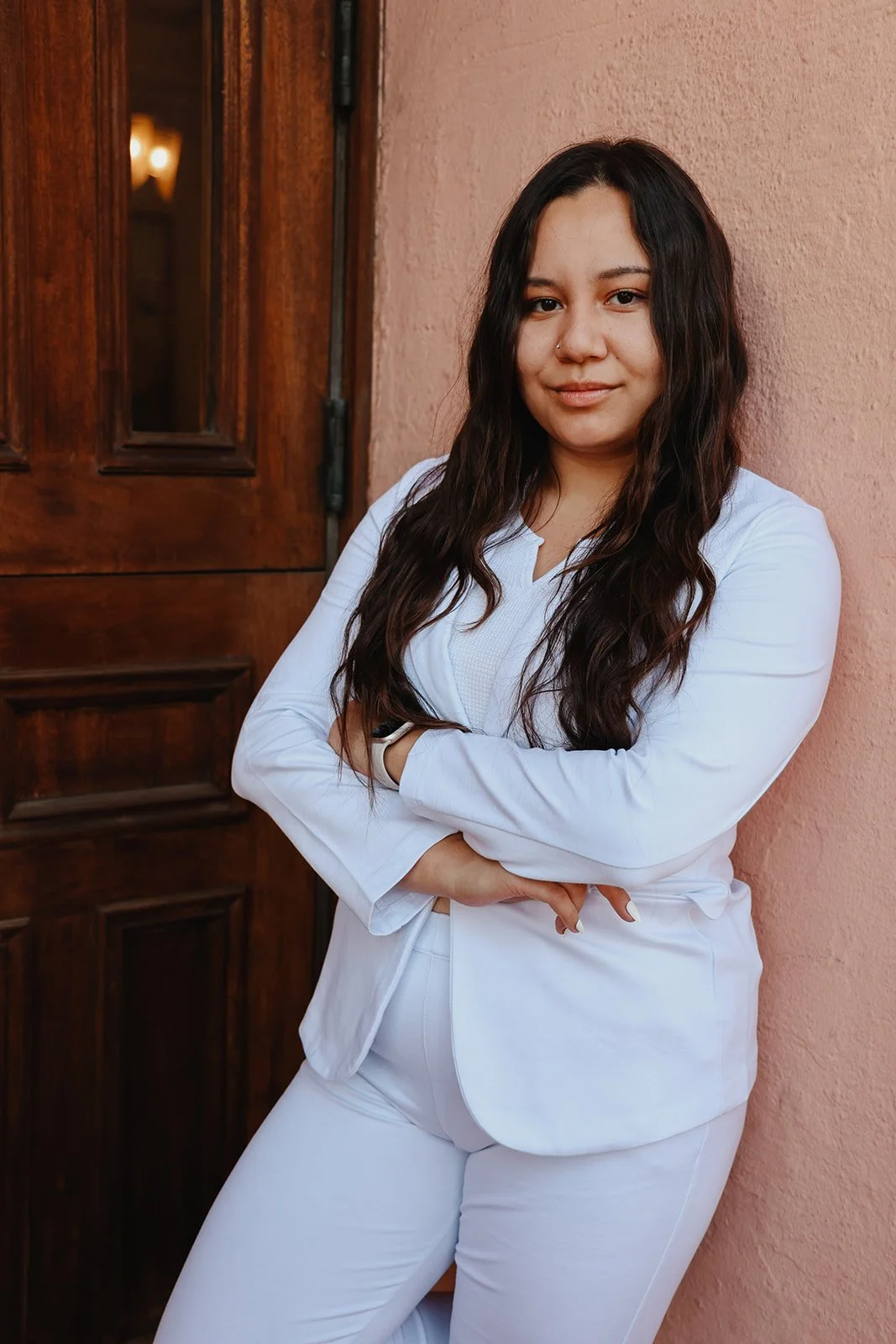 A woman in a white suit leaning against a pink wall with a wooden door in the background.