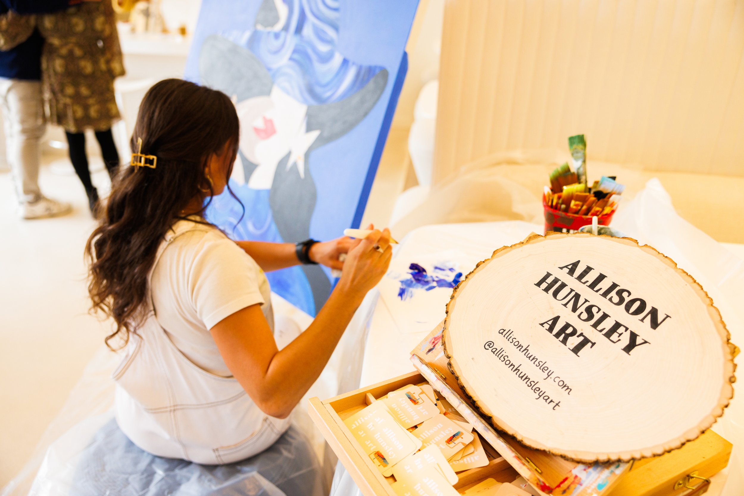 Woman painting on a canvas at an art event, with a large wood sign displaying her name and social media handles in front of her.
