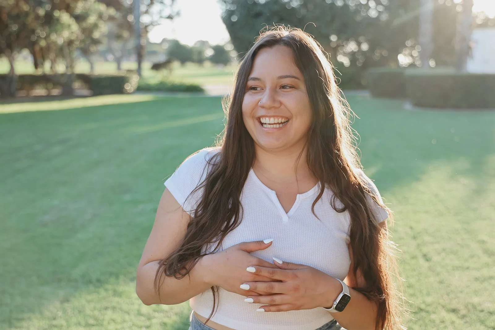 A smiling woman with long dark hair, wearing a white t-shirt and an Apple Watch, standing outdoors in a park with green grass and trees, capturing a joyful moment.