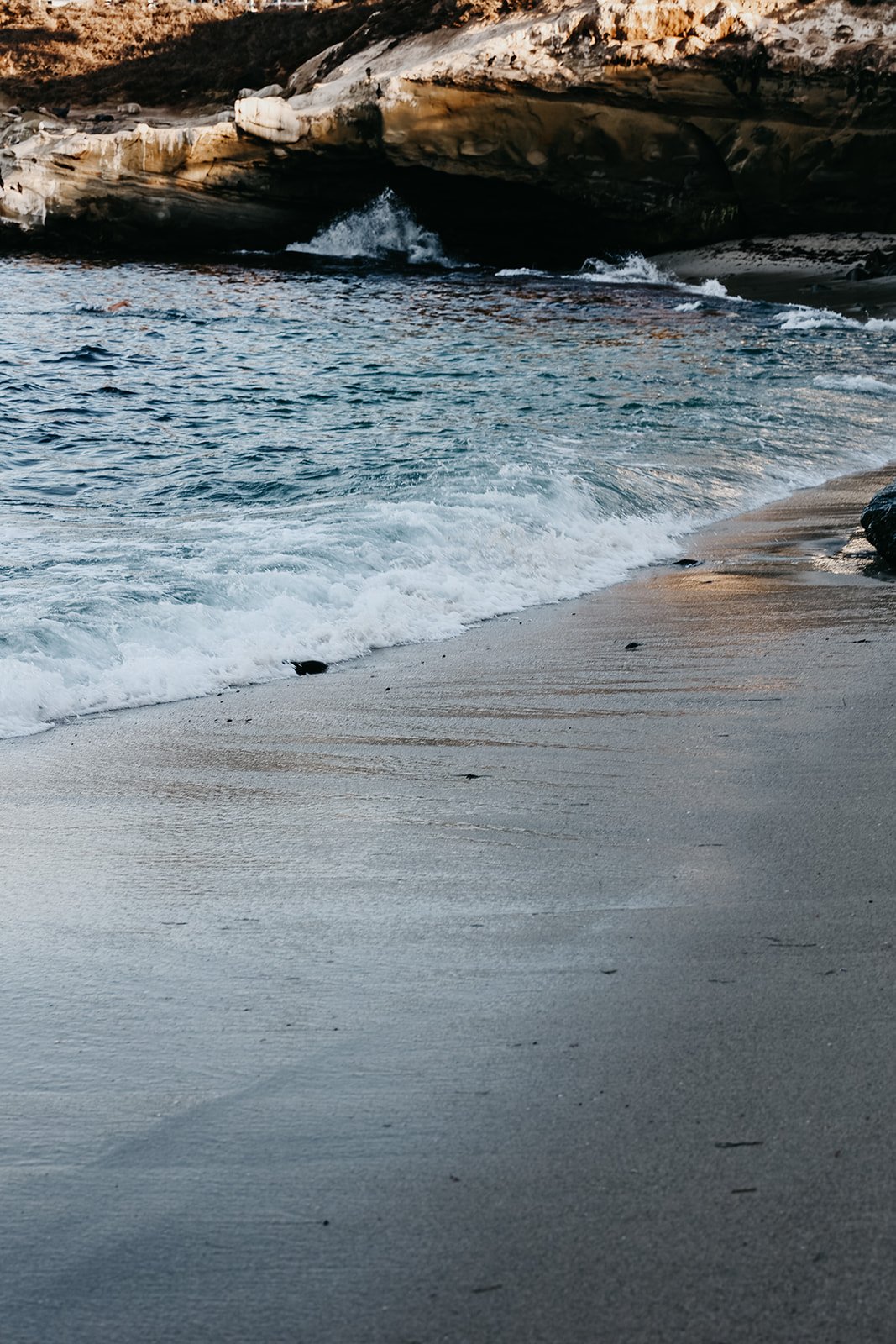 A sandy beach with gentle waves washing onto the shore, with rocky formations in the background.