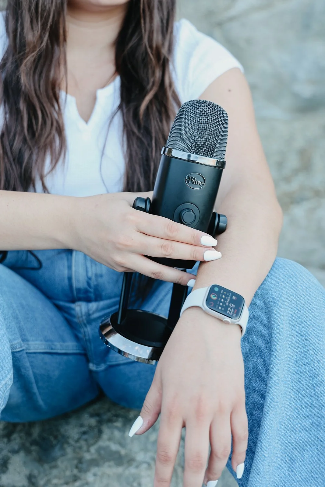 A woman sitting on the ground holding a microphone with her right hand and wearing a smartwatch on her left wrist.