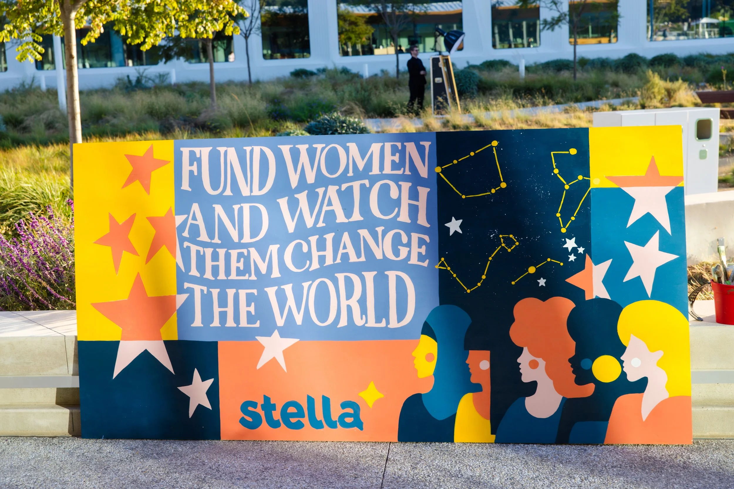 A colorful sign with the message 'Fund Women and Watch Them Change the World' and the name 'Stella.' The sign features illustrations of women, stars, and constellation patterns, set outdoors with a building and trees in the background.