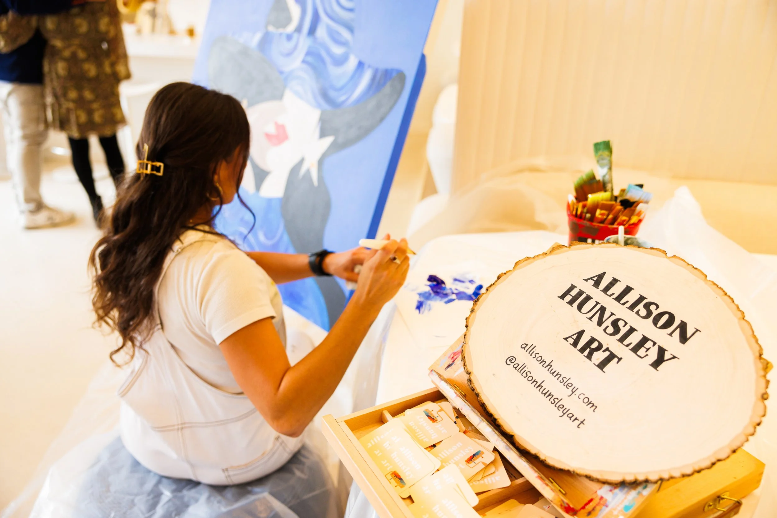 Woman painting at an art exhibition, with a sign reading 'Allison Hunsley Art' and colorful supplies nearby.