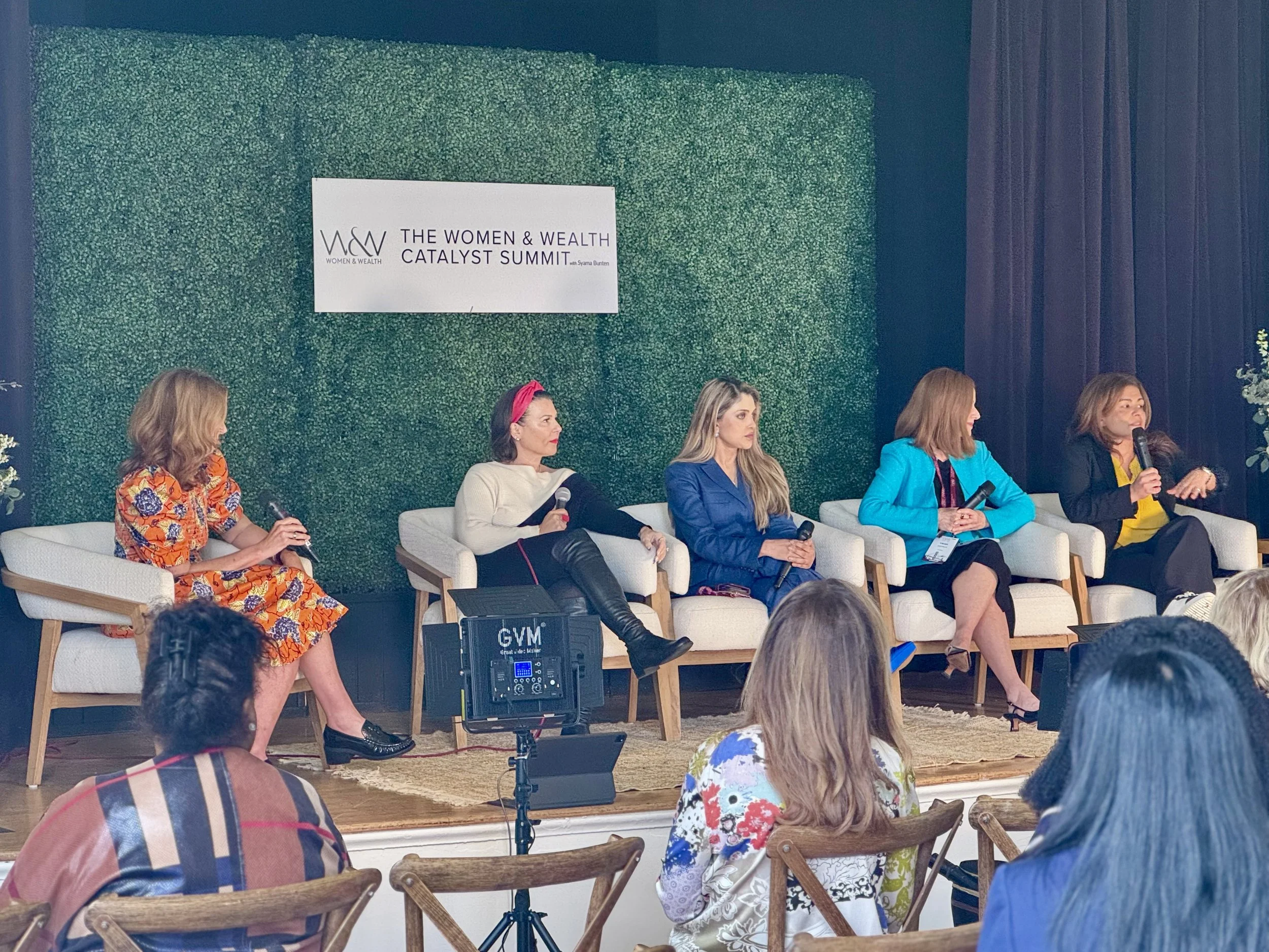 Women participating in panel discussion at 'The Women & Wealth Catalyst Summit' with five women seated on stage, holding microphones, and an audience in front.