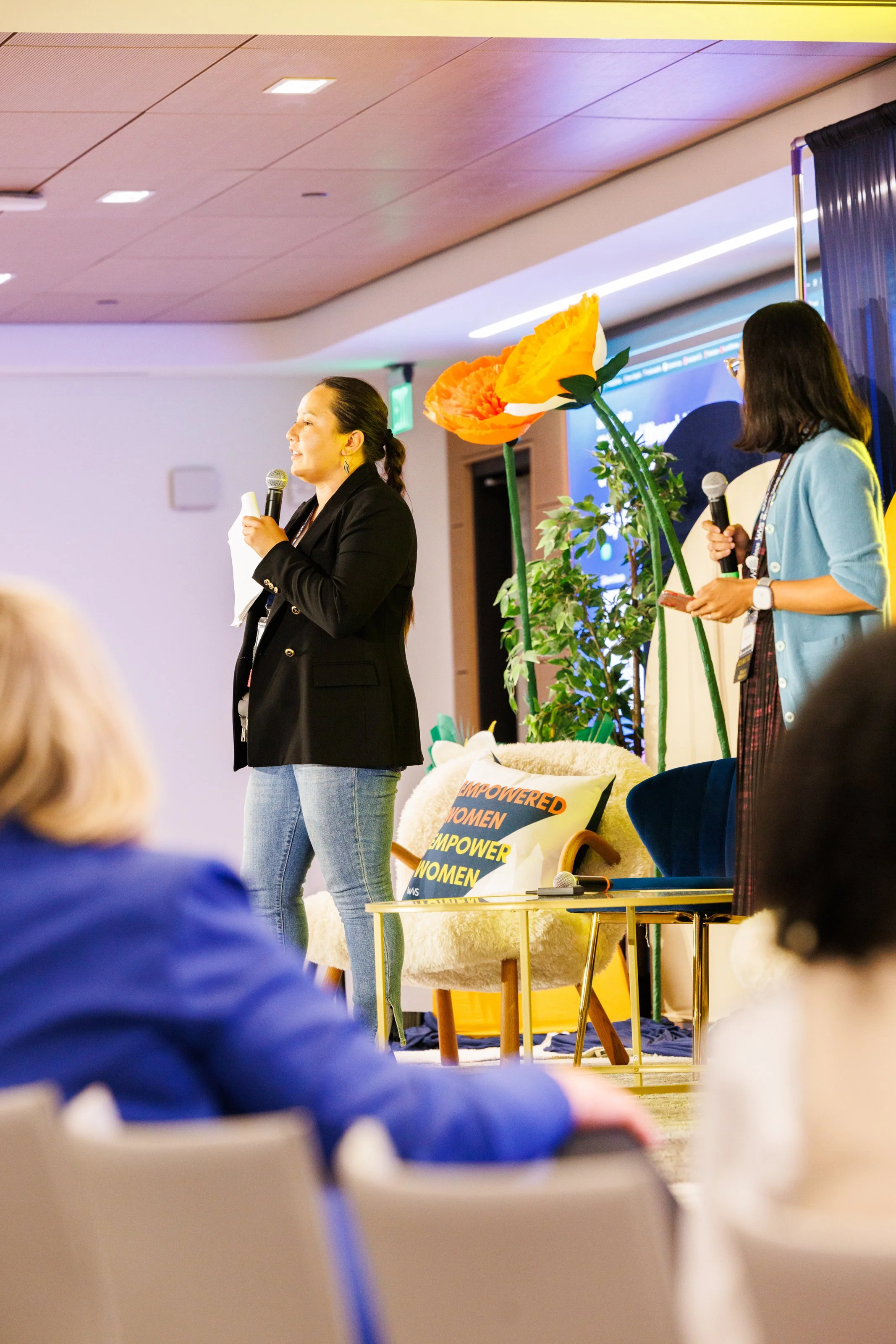 Rylie Jennings speaking at a conference stage with microphones, decorated with large artificial flowers and a pillow that reads 'Empowered Women'.